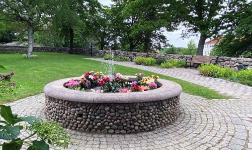 Stensatt fontän med många blomsterbuketter i. I bakgrunden en stenlagd gång, rabatter och bänkar samt mycket grön växtlighet runtom.