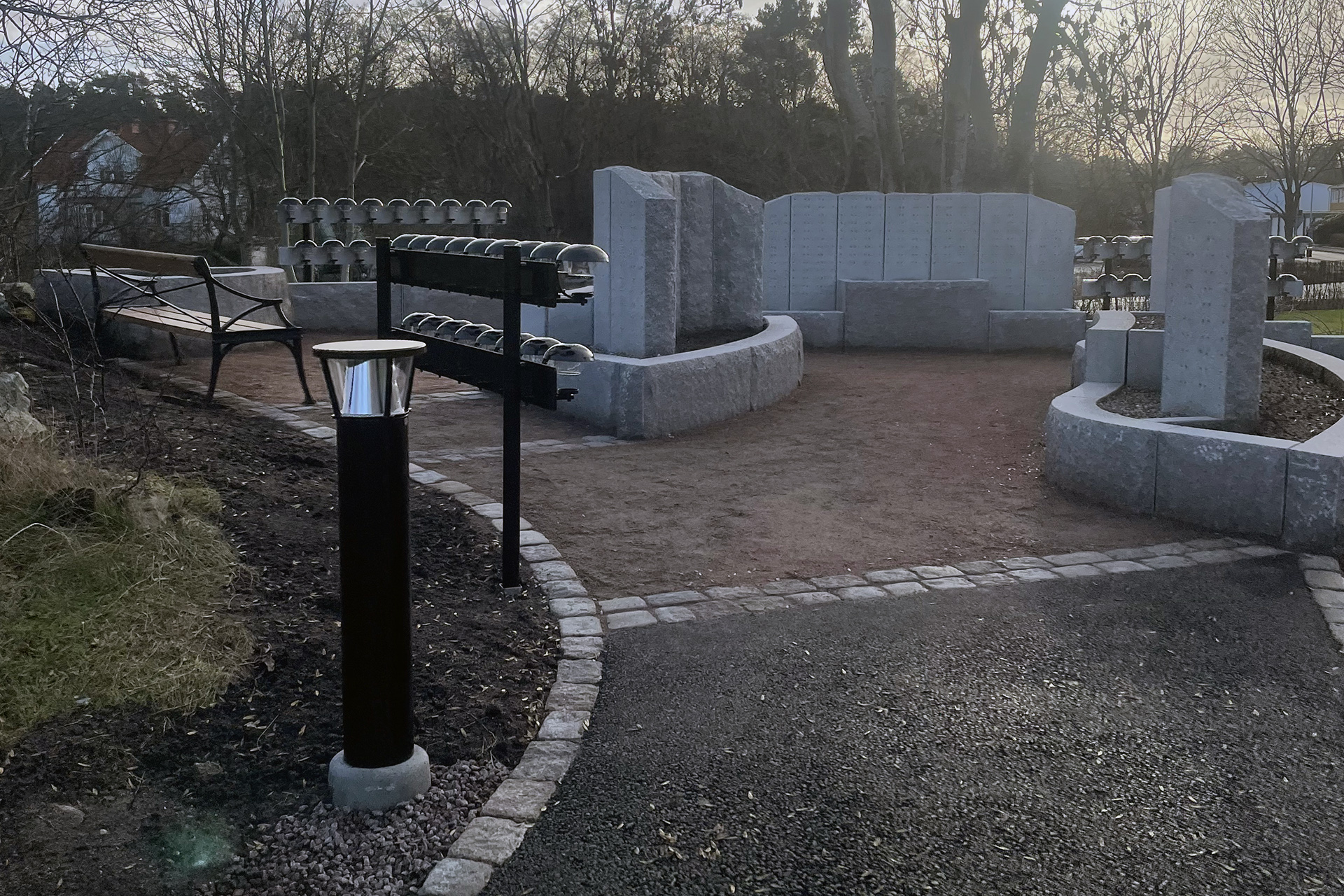 En asfalterad gångväg med stenkant leder fram till en grusad yta med murar och pelare i sten. Vinterkala träd i bakgrunden.