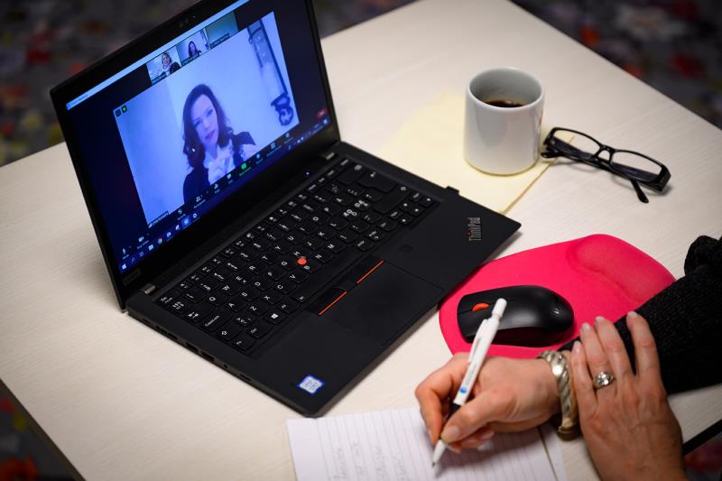 Hand håller i penna intill uppslagen dator med utbildare i bild på skärmen. 