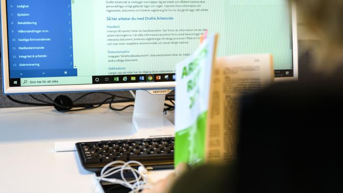 En person sitter intill en datorskärm med en halvt uppslagen bok om arbetsrätt i handen. 