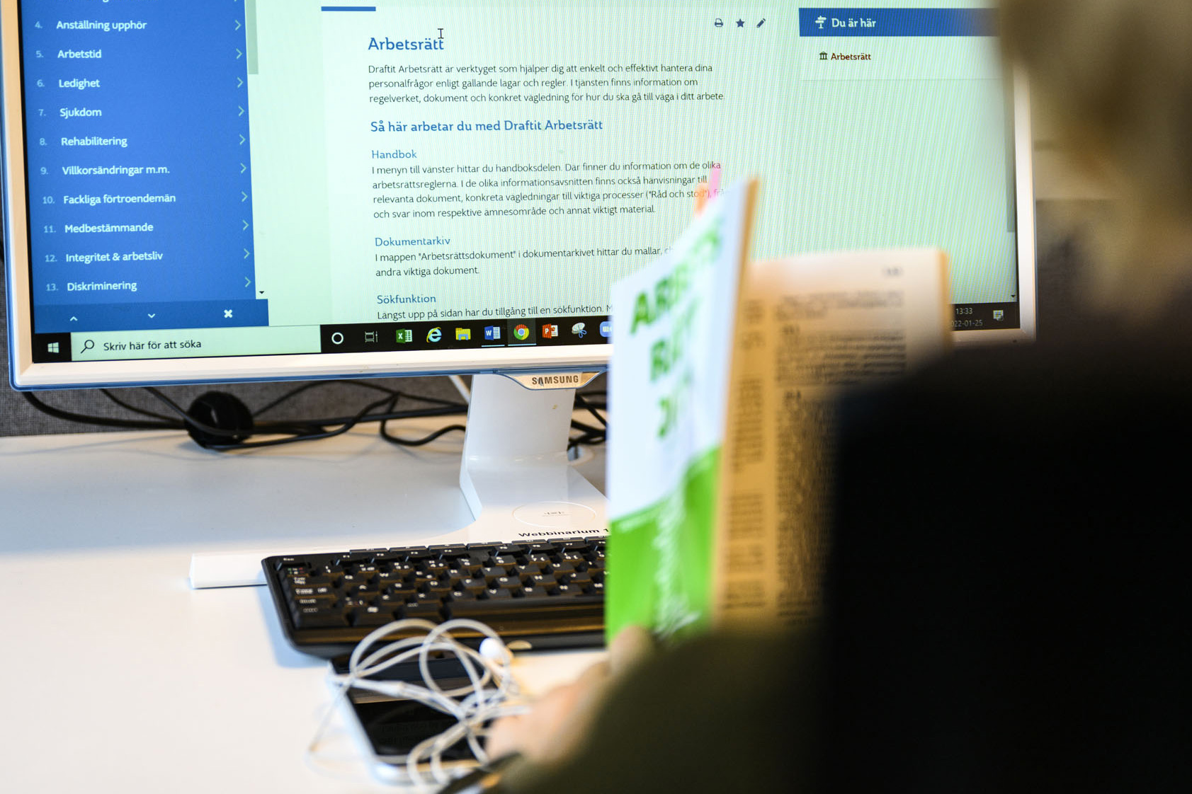 En person sitter intill en datorskärm med en halvt uppslagen bok om arbetsrätt i handen. 