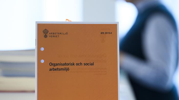 Folder om organisatorisk och social arbetsmiljö i förgrund, person i bakgrund. 