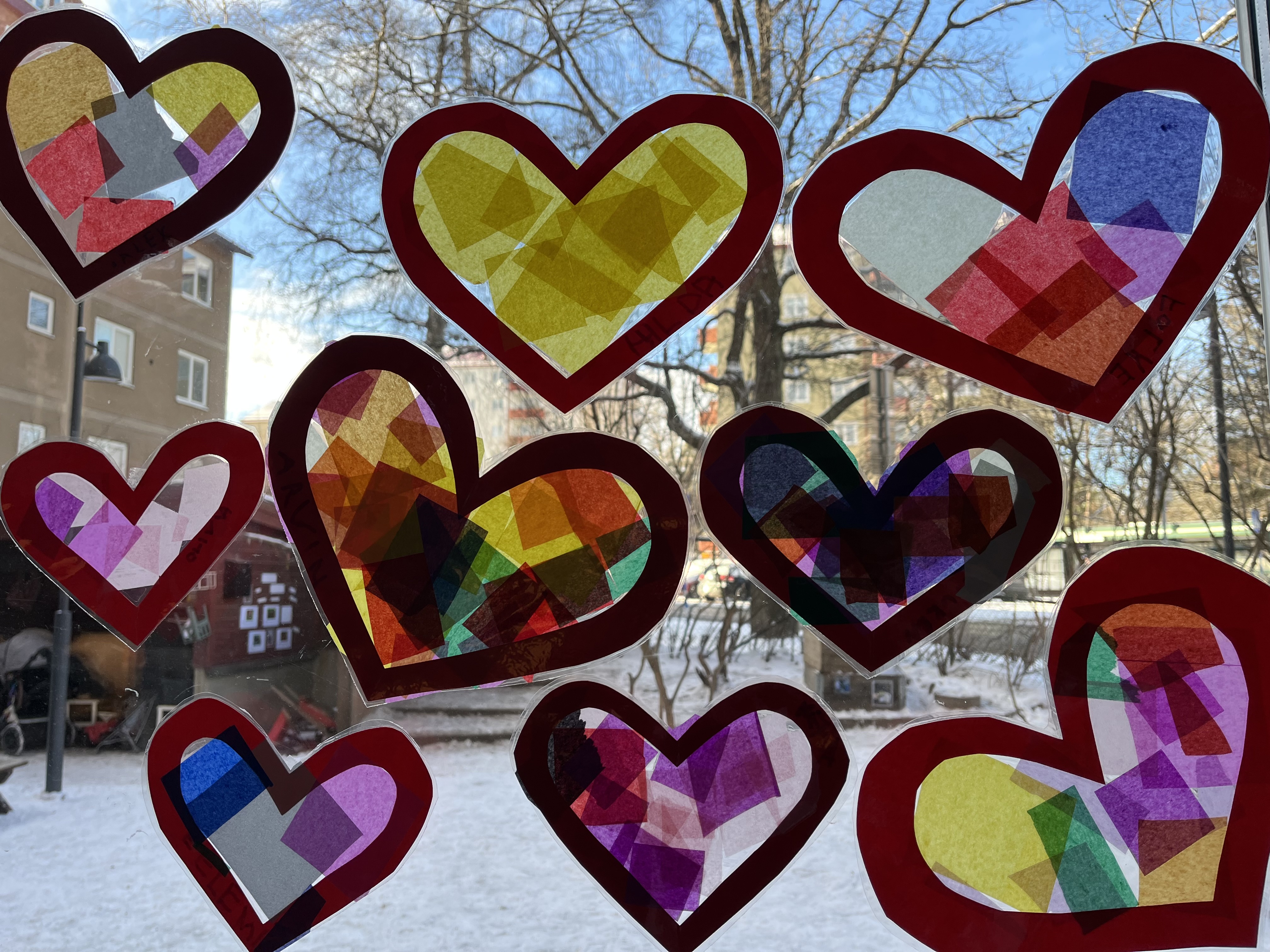Flera färgglada hjärtan gjorda av silkespapper och rött pappersram sitter uppsatta på ett fönster. Solljuset lyser igenom och skapar en vacker effekt. Utanför syns en snötäckt gård med träd och byggnader i bakgrunden.