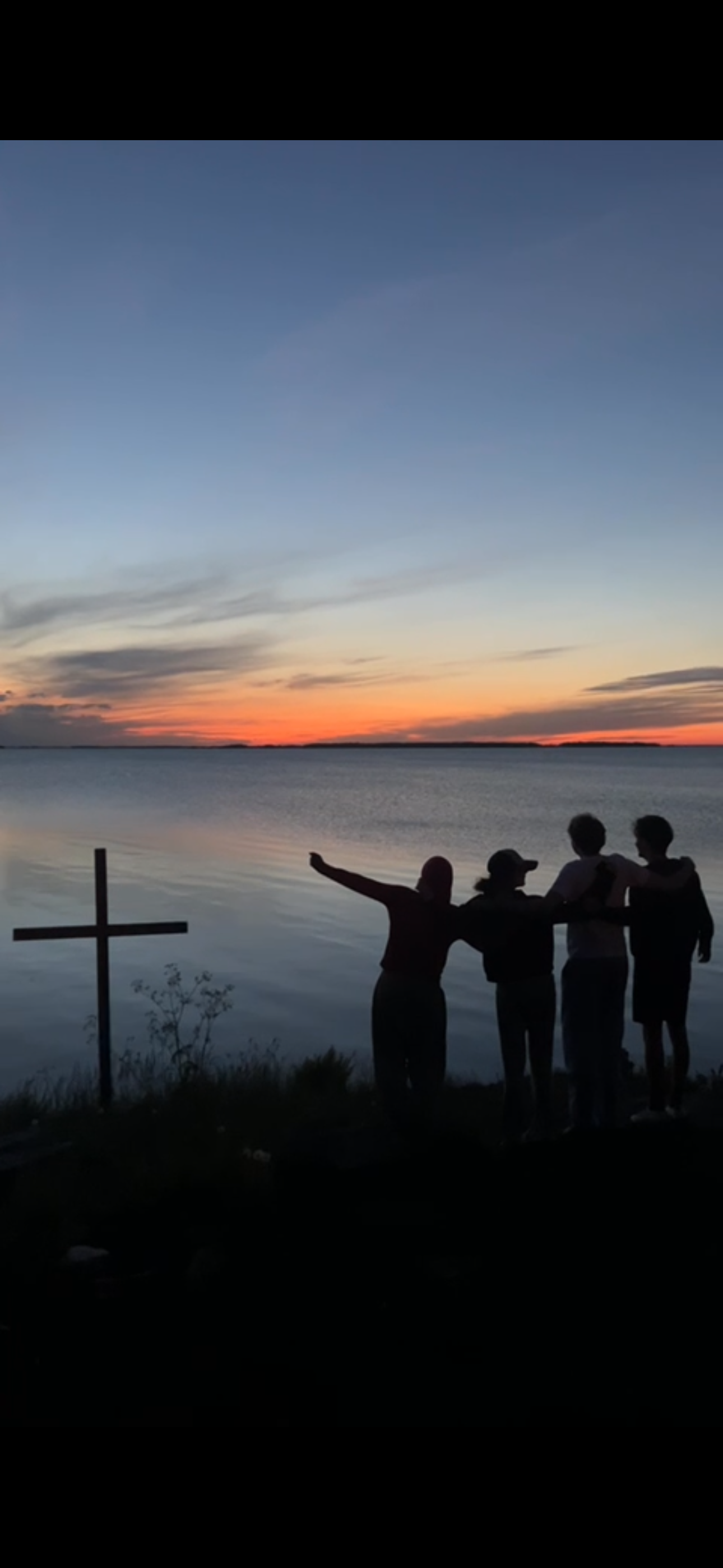 Siluett av fyra ungdomar framför ett kors och en solnedgång vid vattnet.