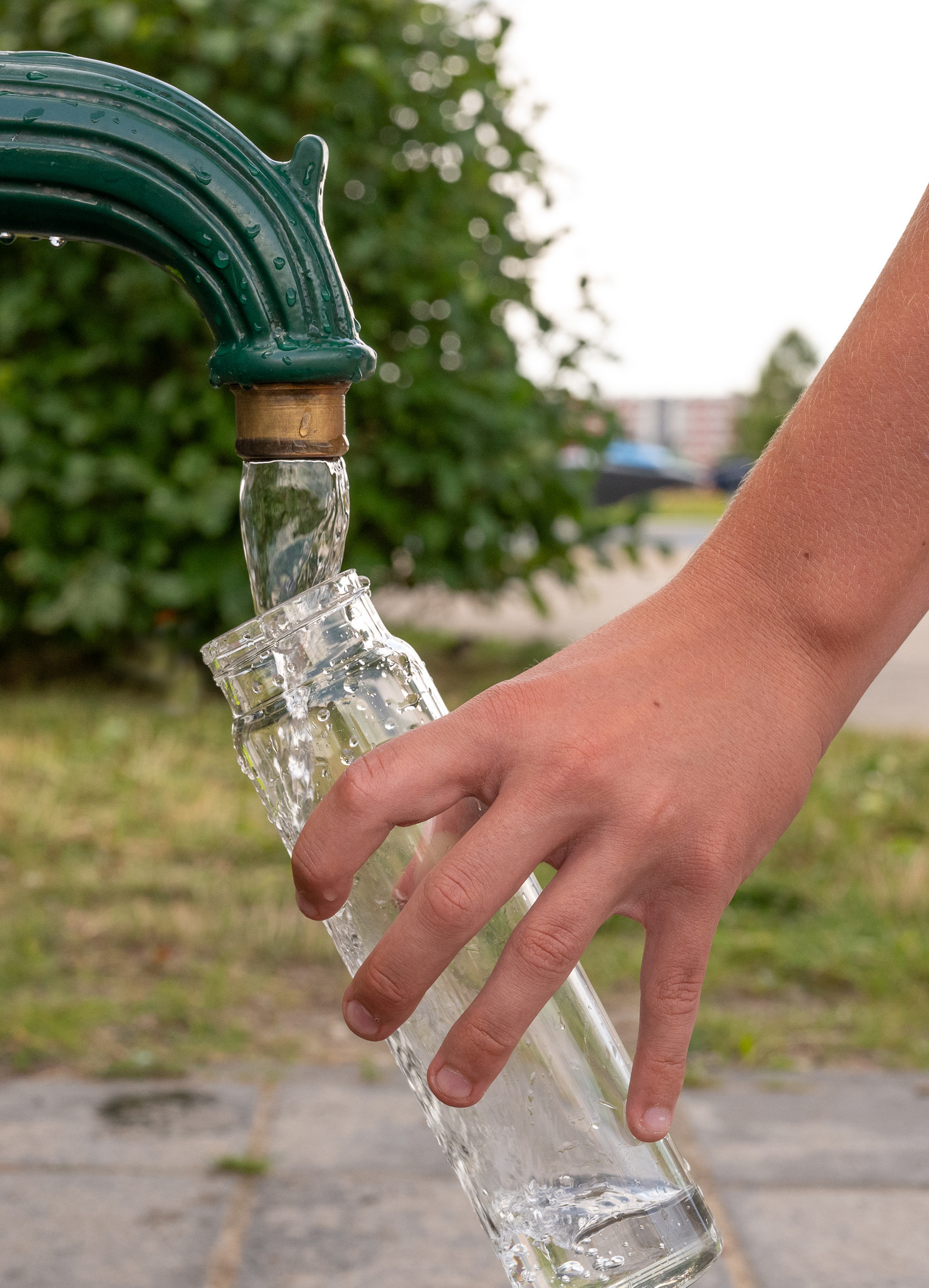 En hand håller en lutad glasflaska under en grön utomhuskran medan klart vatten rinner ner i flaskan. I bakgrunden syns grönska och en suddig utomhusmiljö.