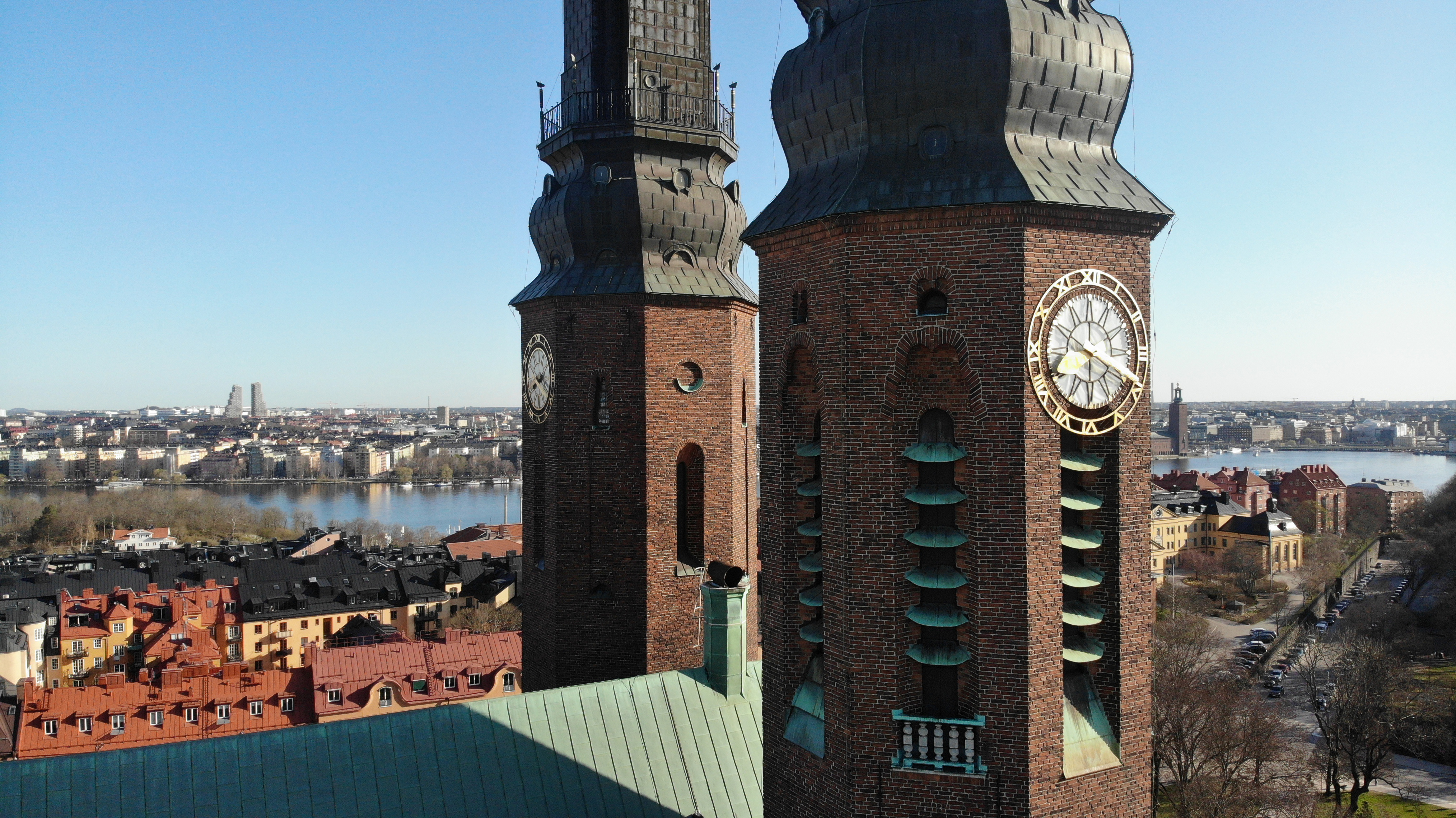 Flygfoto av Högalidskyrkans två tegelklädda kyrktorn med stora urtavlor. I bakgrunden syns Stockholms stadssiluett, vatten och bebyggelse under en klarblå himmel.