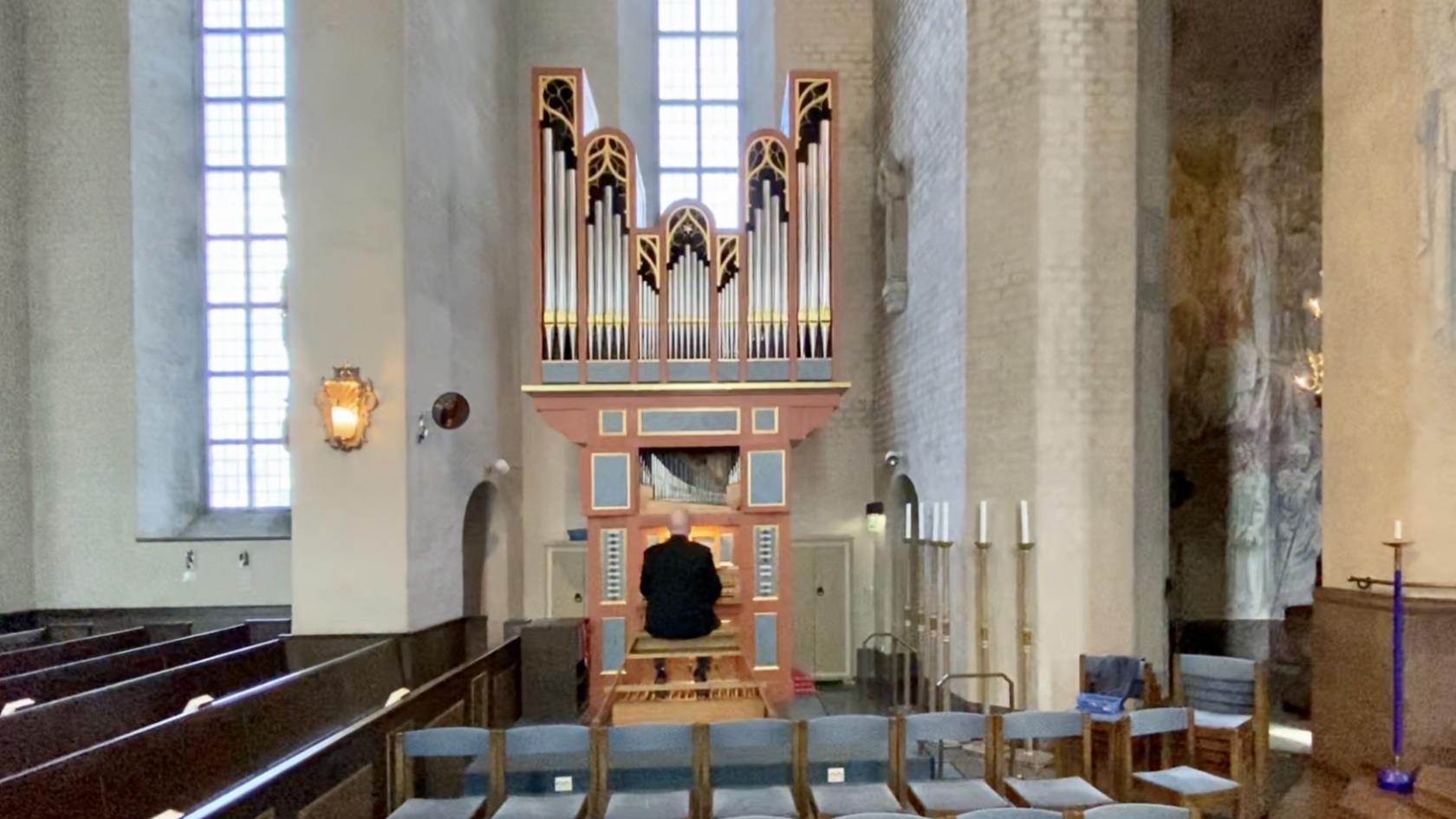 En orgel och en person som sitter vid den. kyrkbänkar, ljus, höga fönster. 