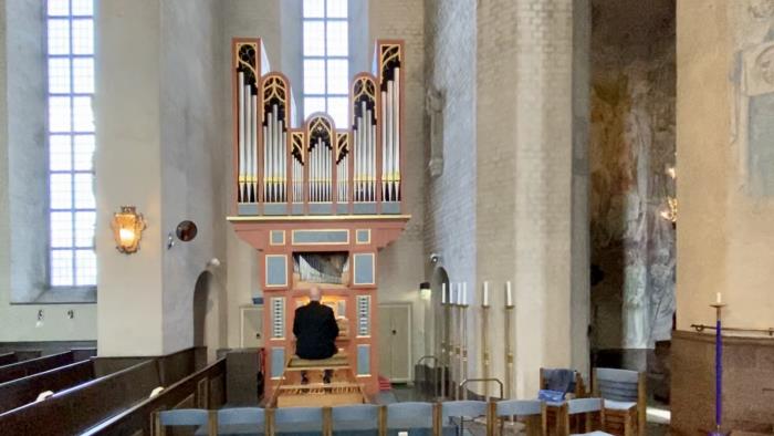 En orgel och en person som sitter vid den. kyrkbänkar, ljus, höga fönster. 