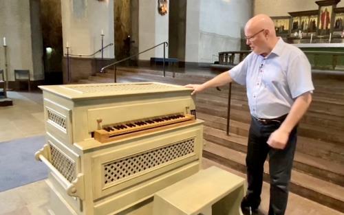 En liten beigefärgad orgel.  En man står bredvid och håller ena handen på orgeln. 