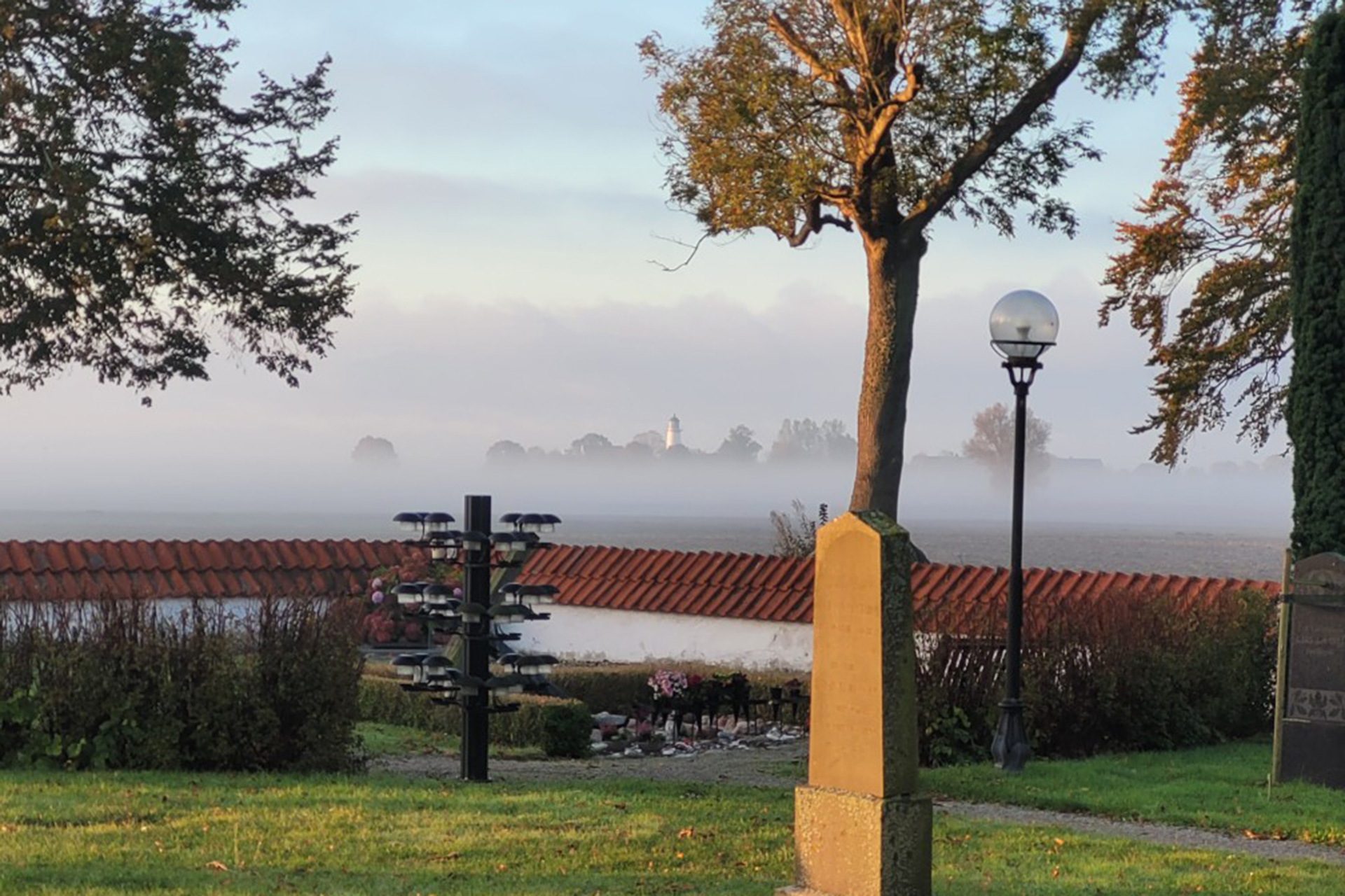 En gravsten och en ljusbärare står på en kyrkogård med utsikt över havet. I fjärran syns en fyr. Över havet ligger en dimslöja.