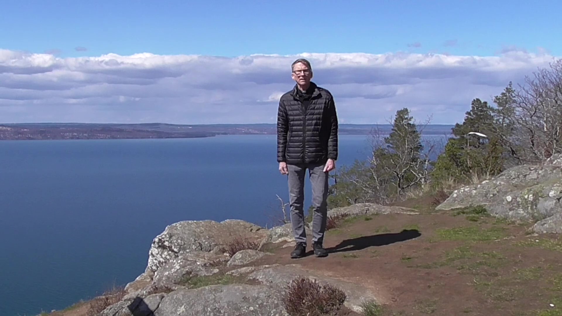 Prästen Ingemar Persson står på Brunstorpsberget med ett blått glittrande vättern i bakgrunden.
