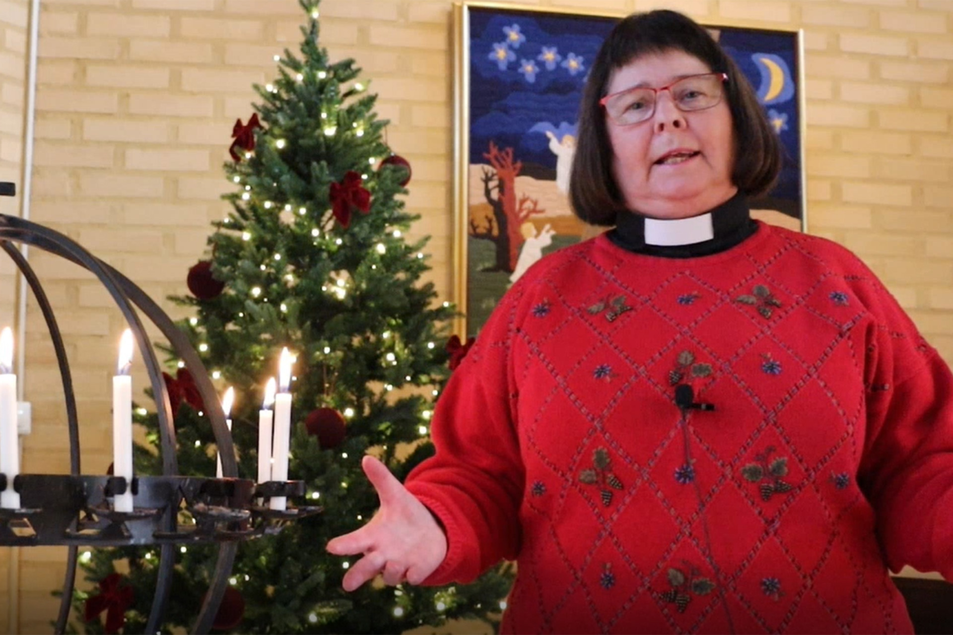 En kvinnlig präst i prästskjorta och röd tröja står bredvid en ljusbärare och pratar