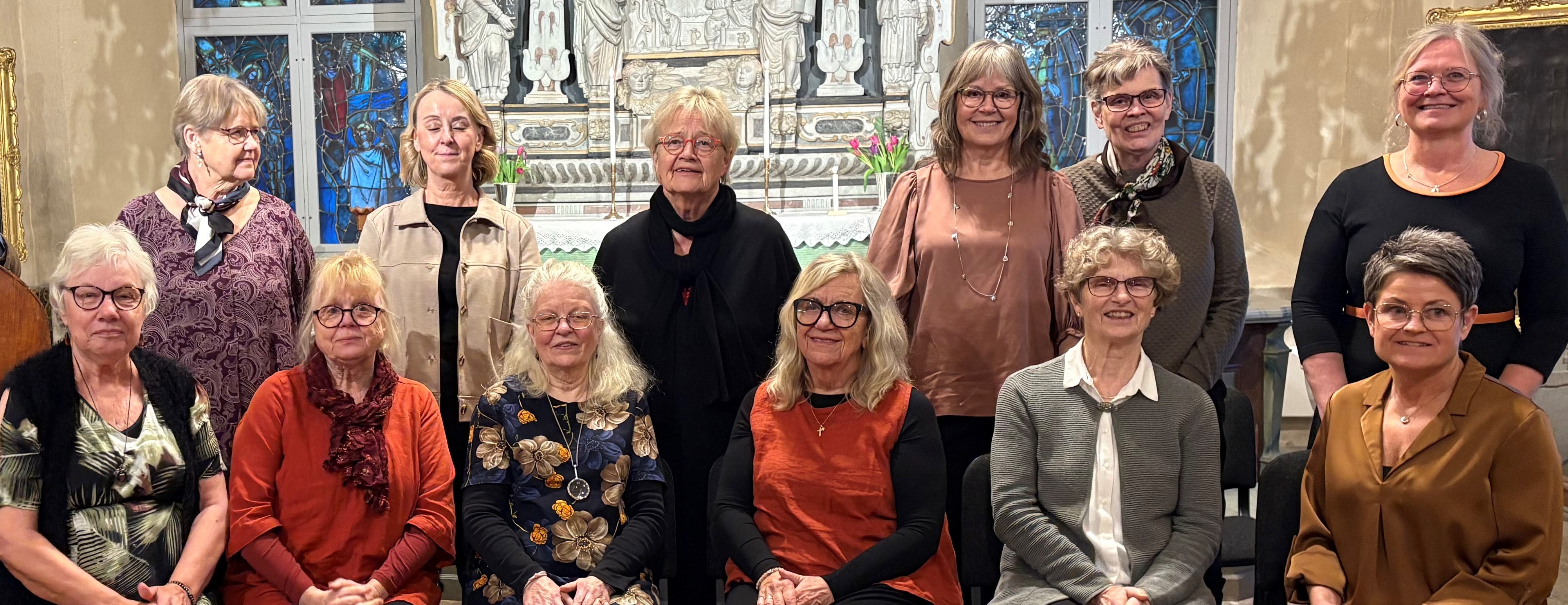 Körmedlemmar som sitter och står tillsammans med ledaren Maria Norgen framför altaret i Rasbo kyrka