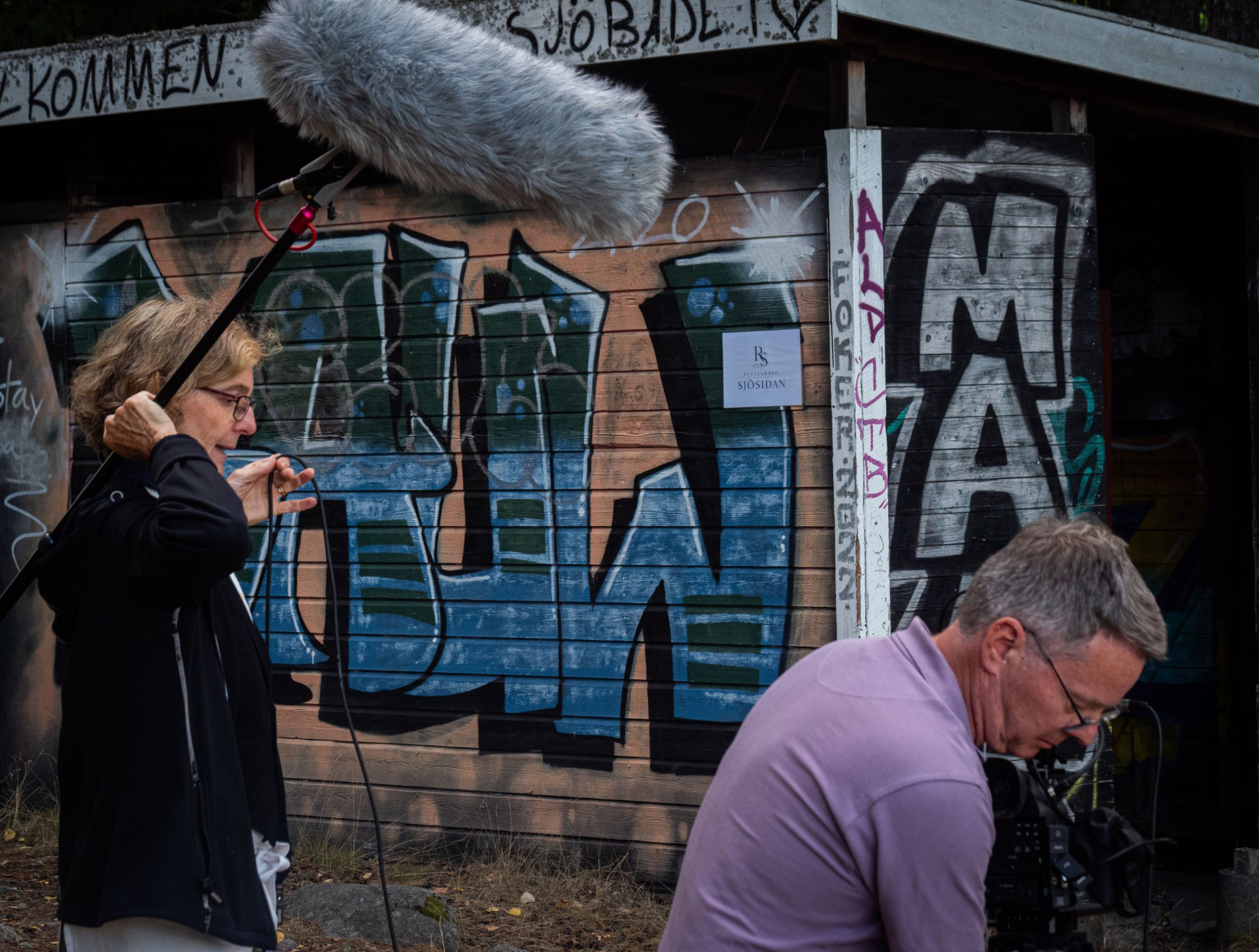 Kvinna står med stor mikrofon med ludd på och man lutar sig framåt mot filmkamera utomhus med grafittivägg bakom.
