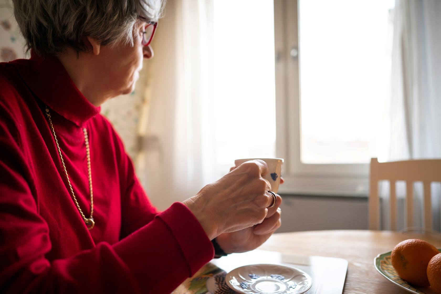 Bild på kvinna som sitter med kaffekopp och tittar ut genom fönstret och ser ensam, ledsen ut