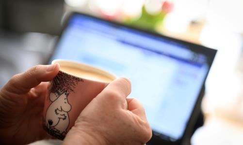 Händer håller i kaffemugg. Datorskärm i bakgrunden