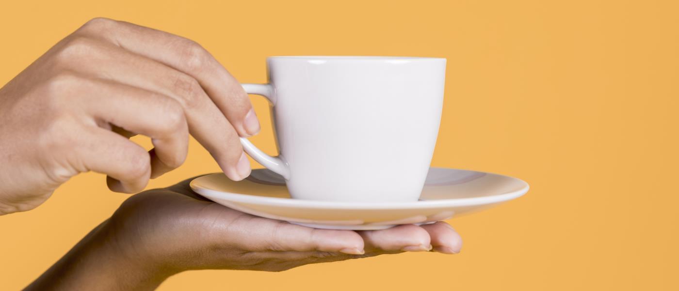 Ett par händer håller fram en vit kaffekopp med fat. Bakgrunden är orange och enfärgad