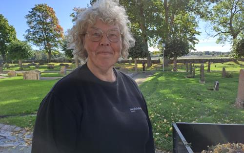 En kvinna med kort lockigt, grått hår och glasögon tittar leende in i kameran. Hon står på en kyrkogård med grönt gräs.