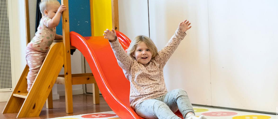 Att barn sitter vid en rutchbana och sträcker upp armarba och ser glad ut. Ett annat barn står på stegen på rutchbanan.