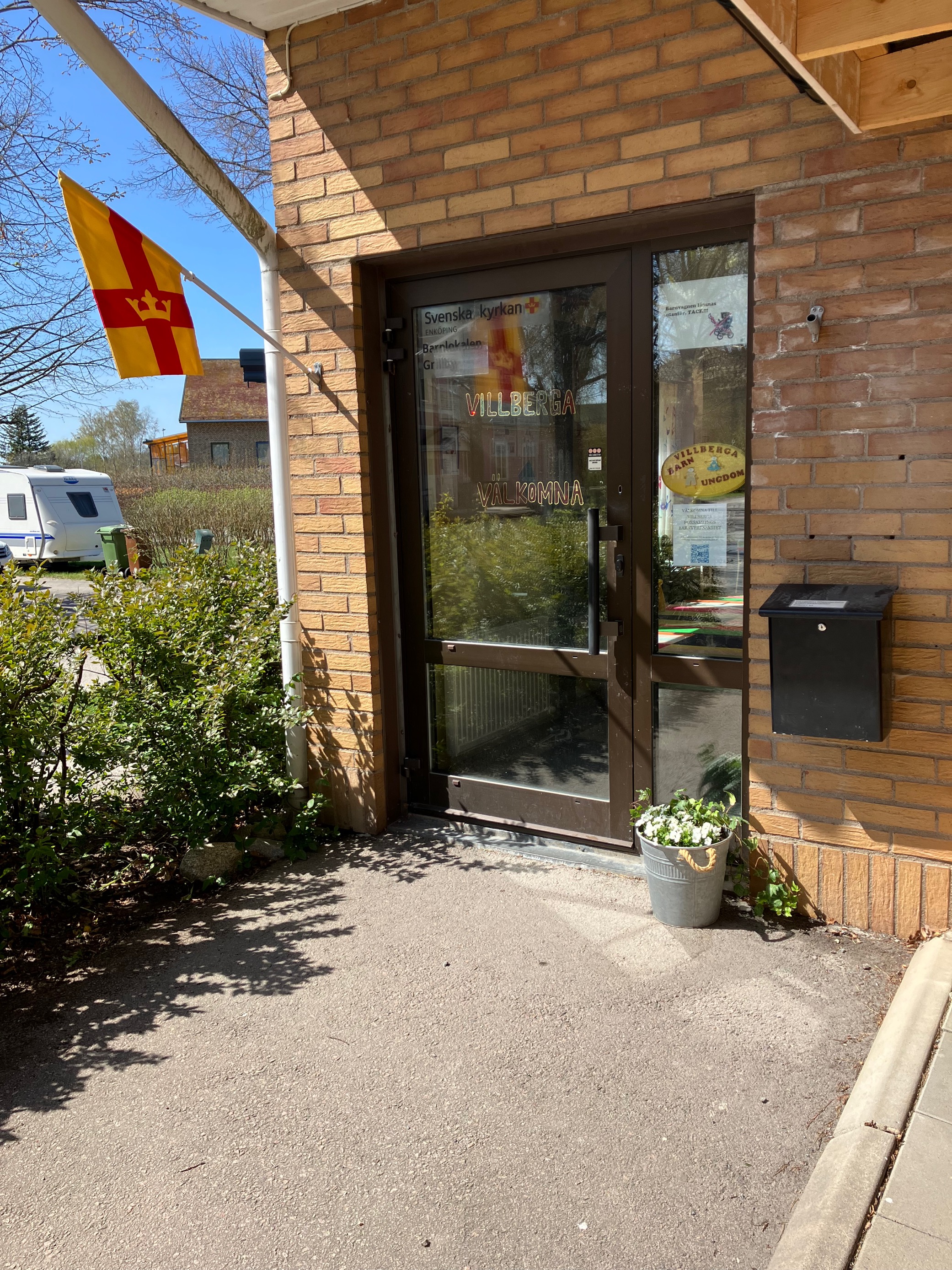 En entrédörr till en gul tegelbyggnad. Bredvid dörren finns en häck, lite växter och Svenska kyrkans flagga.
