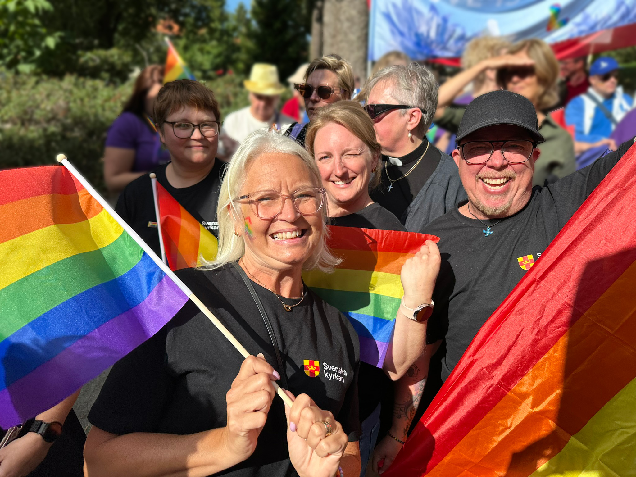 En grupp människor i olika åldrar och av olika kön ler stort mot kameran. De har kläder med Svenska kyrkans logga på och håller i regnbågsflaggor.