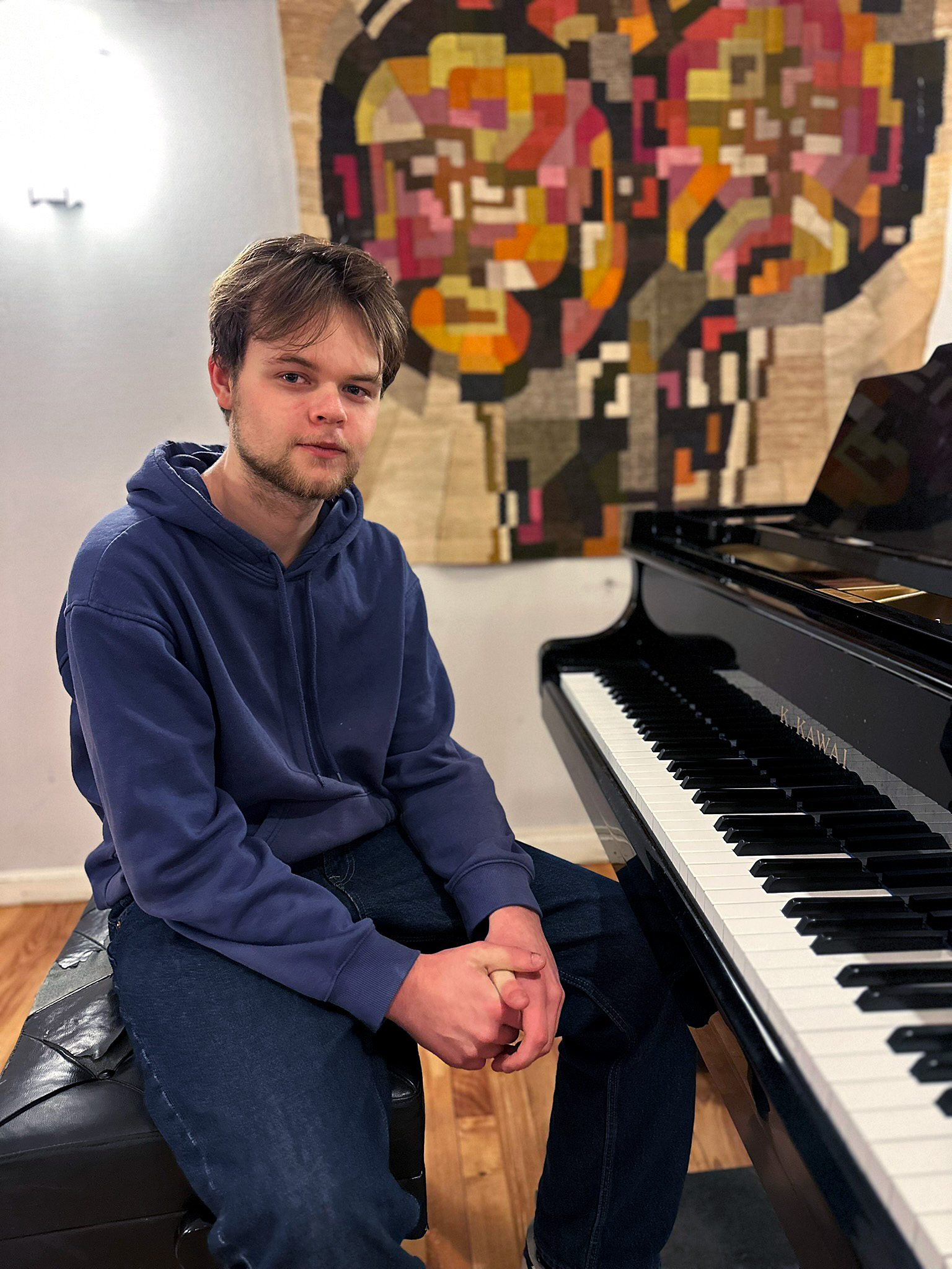 En kille i övre tonåren sitter vid en flygel. han tittar försiktigt in i kameran.