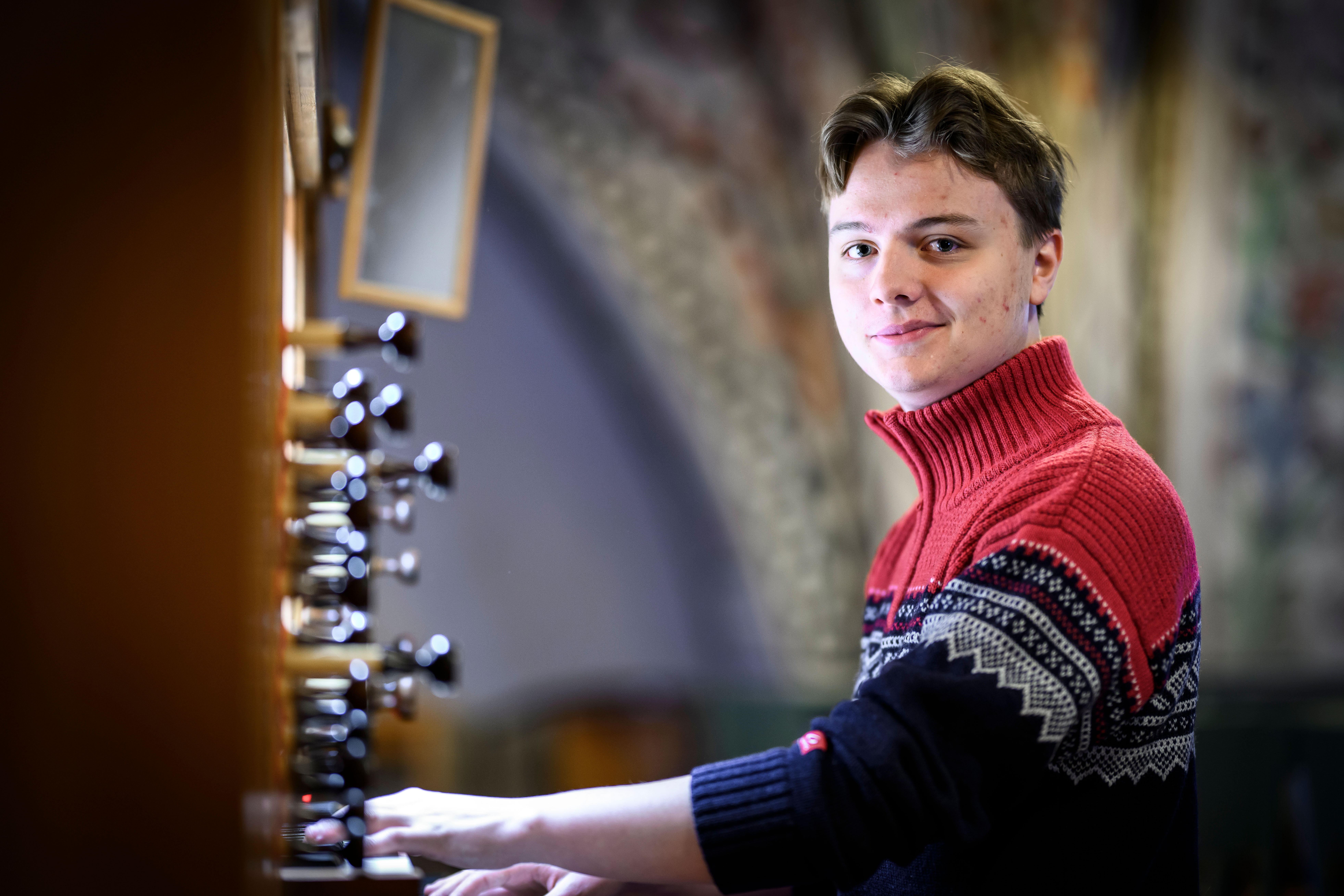 En kille i övre tonåren sitter vid en kyrkorgel och spelar. han tittar in i kameran och ler lite snett.