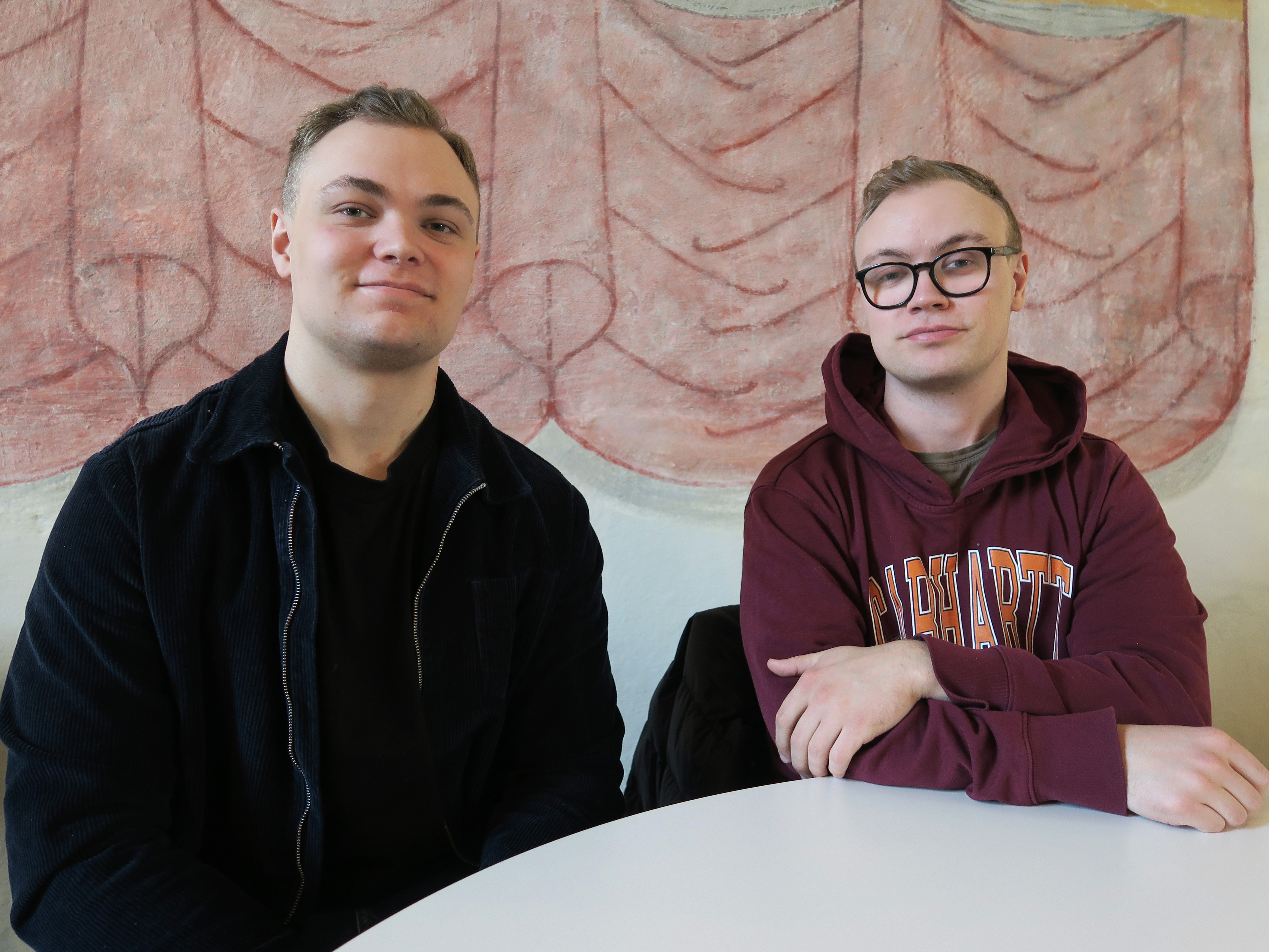 Två killar i tjugoårsåldern sitter vid ett bord. En av dem har svarta kläder och kort hår. Den andra har glasögon och en vinröd hoodie med tryck.