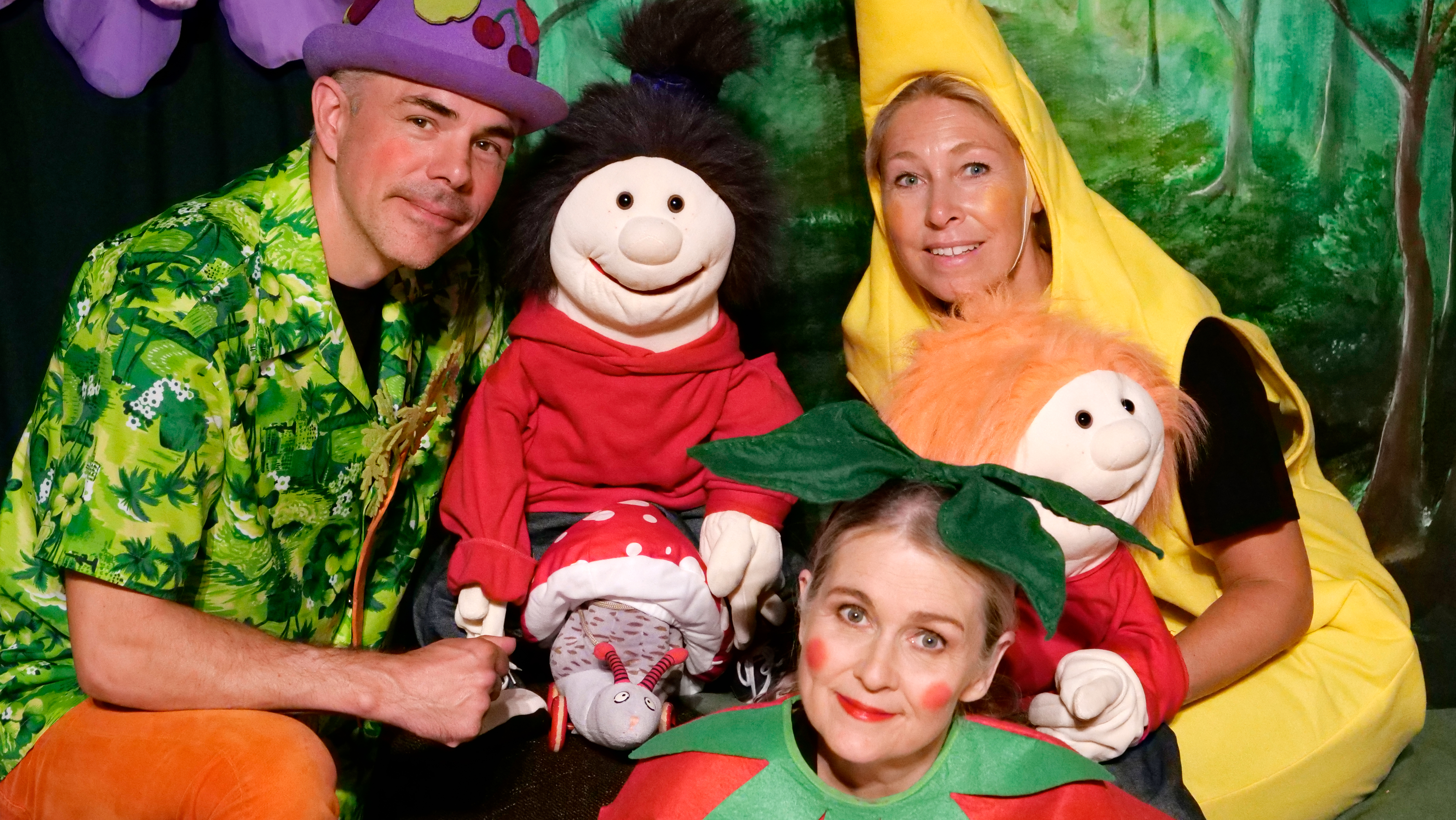 Tre skådespelare utklädda till frukter och två dockor sitter tillsammans på ett golv.