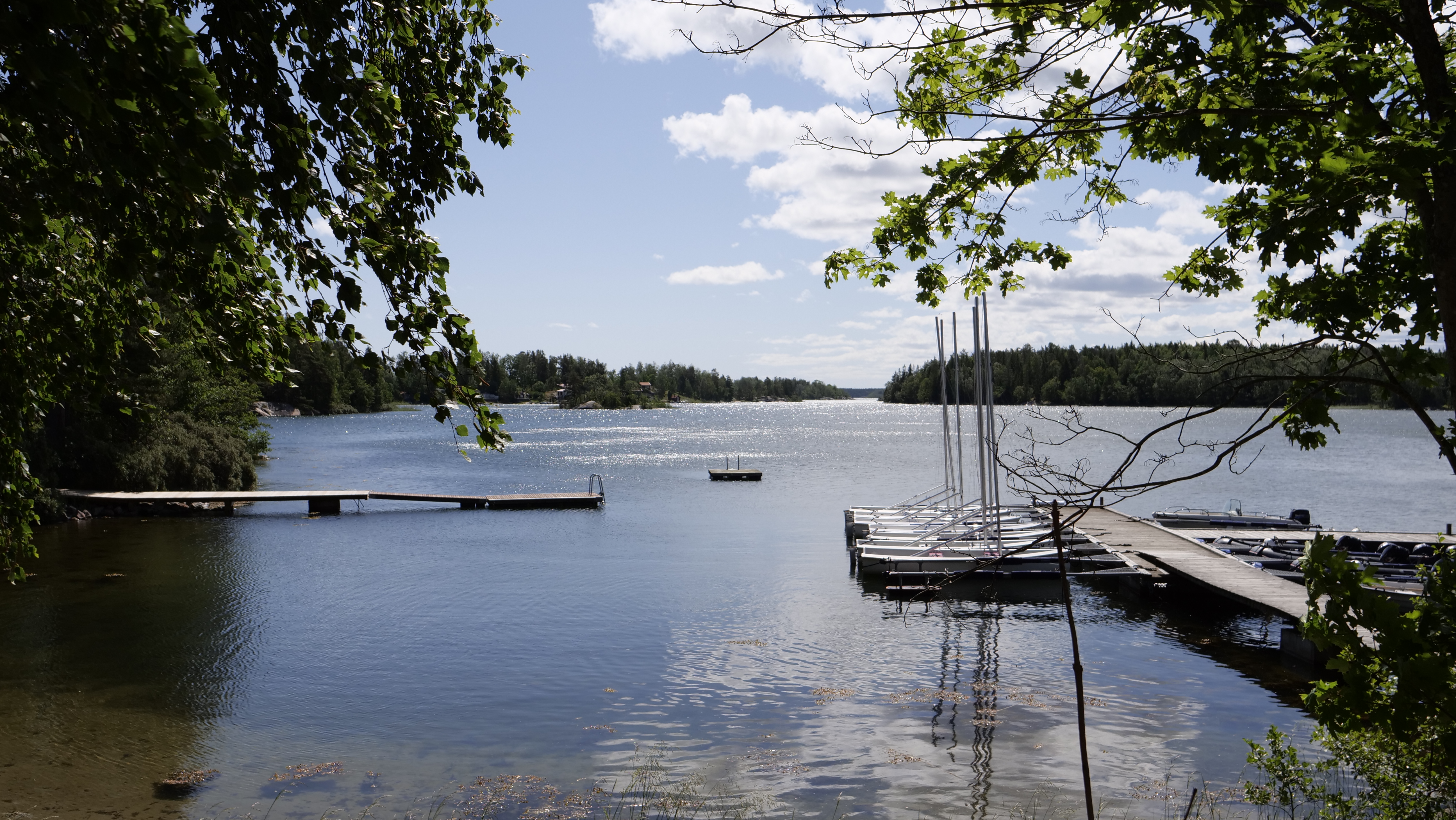 Lägergården Karlberg vid vattnet - bryggor och båtar i vattnet med grönska runt omkring.