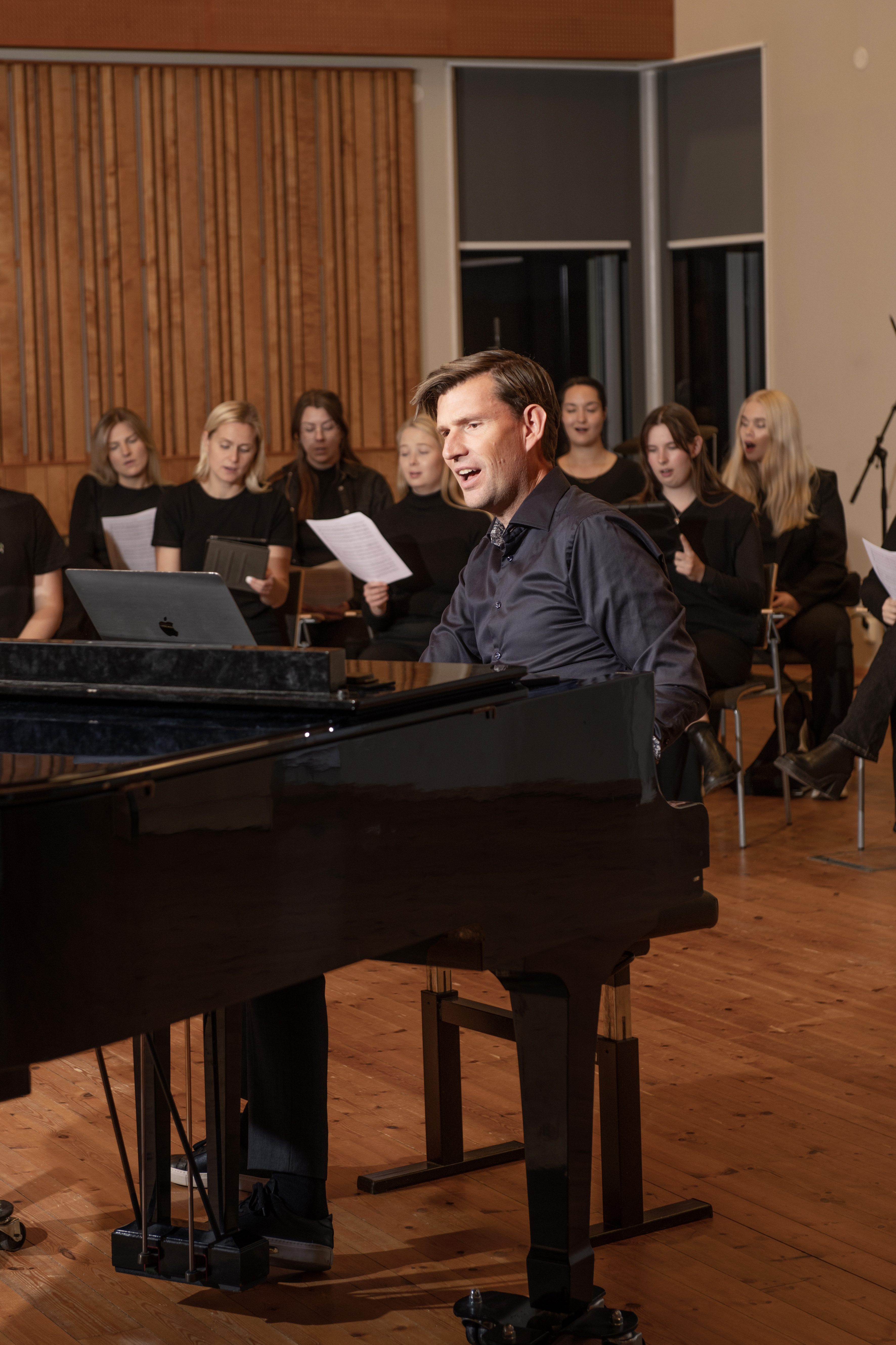 Man sitter vid piano och sjunger. Runt honom sitter personer som också sjunger.