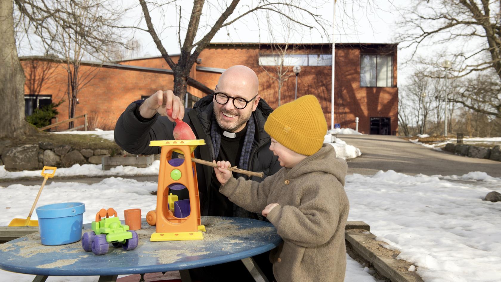 Bilden visar Adolfsbergs kyrka i bakgrunden. I förgrunden syns en präst och ett barn som leker vid lekplatsen.