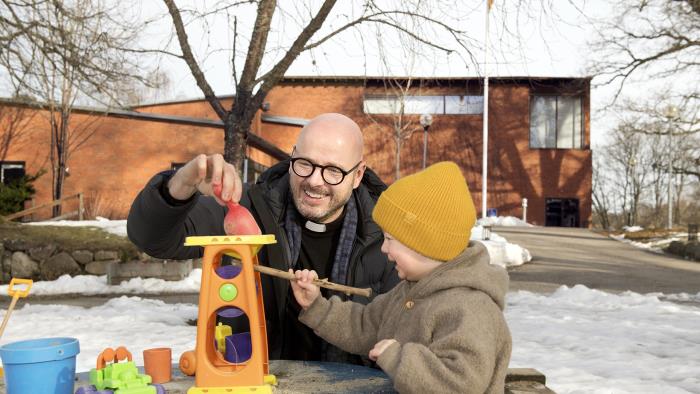 Bilden visar Adolfsbergs kyrka i bakgrunden. I förgrunden syns en präst och ett barn som leker vid lekplatsen.