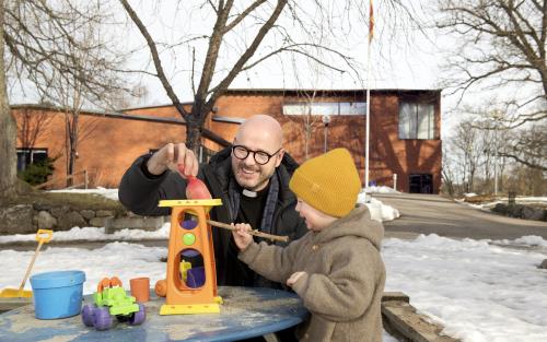 Bilden visar Adolfsbergs kyrka i bakgrunden. I förgrunden syns en präst och ett barn som leker vid lekplatsen.