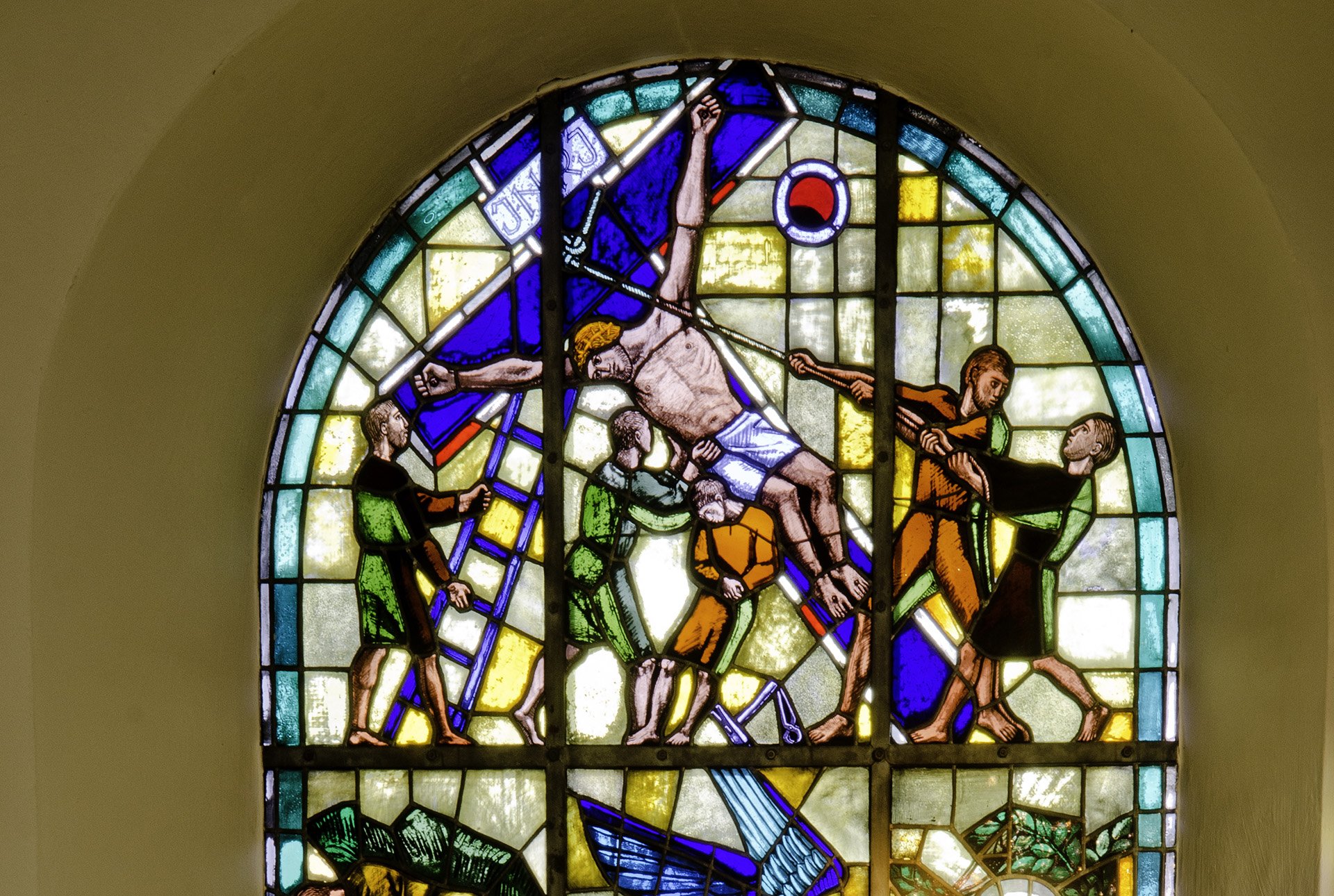 Ett fönster med blyinfattat glas. Det föreställer en scen där Jesus är fastspikad på ett kors som sex män hjälps åt att ställa upp. 