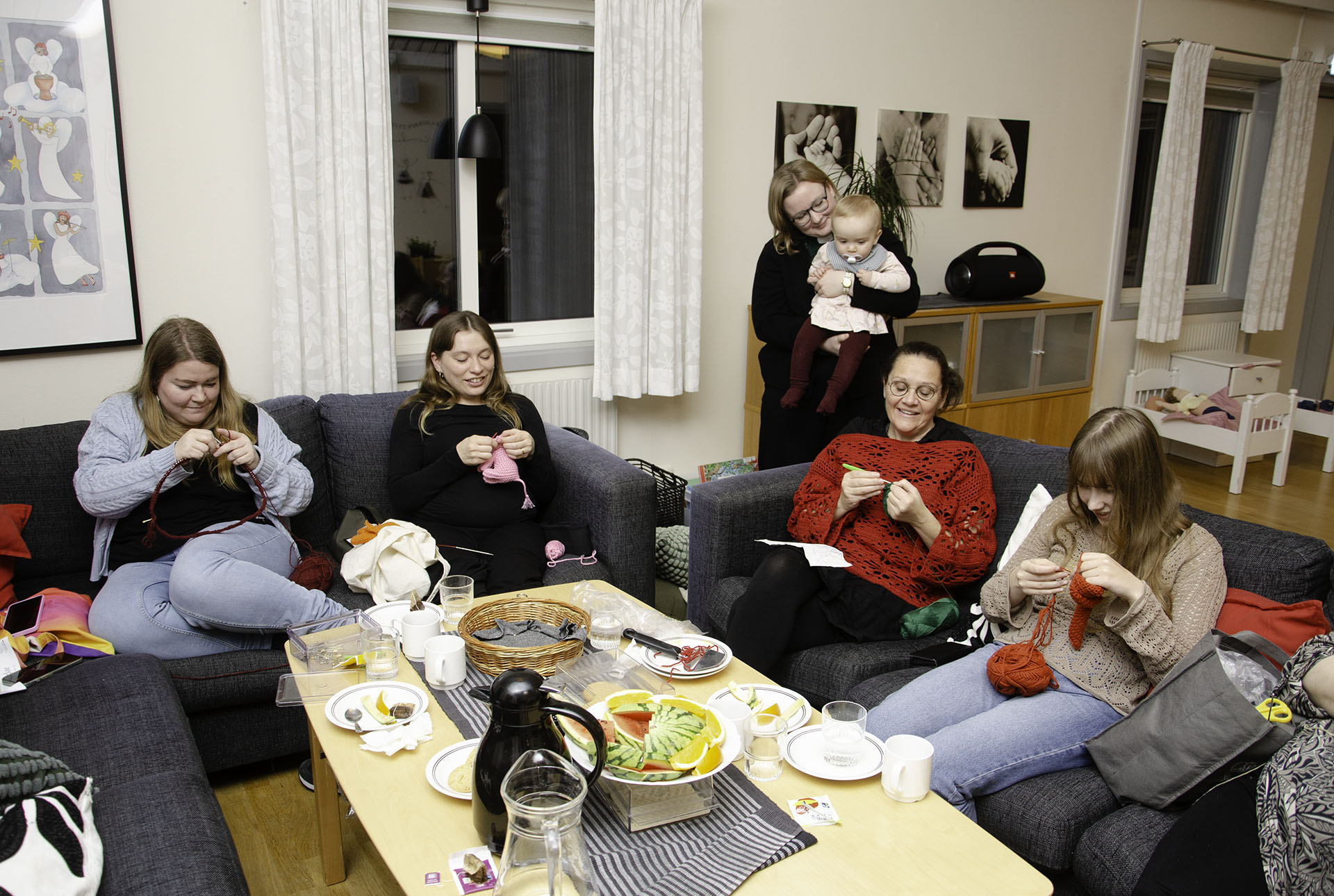 Fyra personer sitter i en soffa och stickar och virkar. En person står och håller ett barn i famnen bakom soffan 
