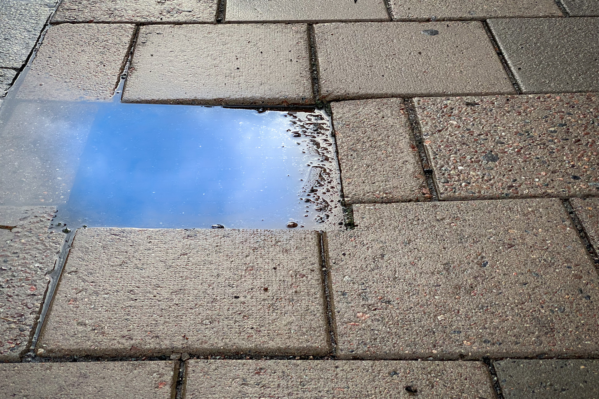 Fotot visar en regnvåt trottoar med en liten vattenpöl som speglar himlen.