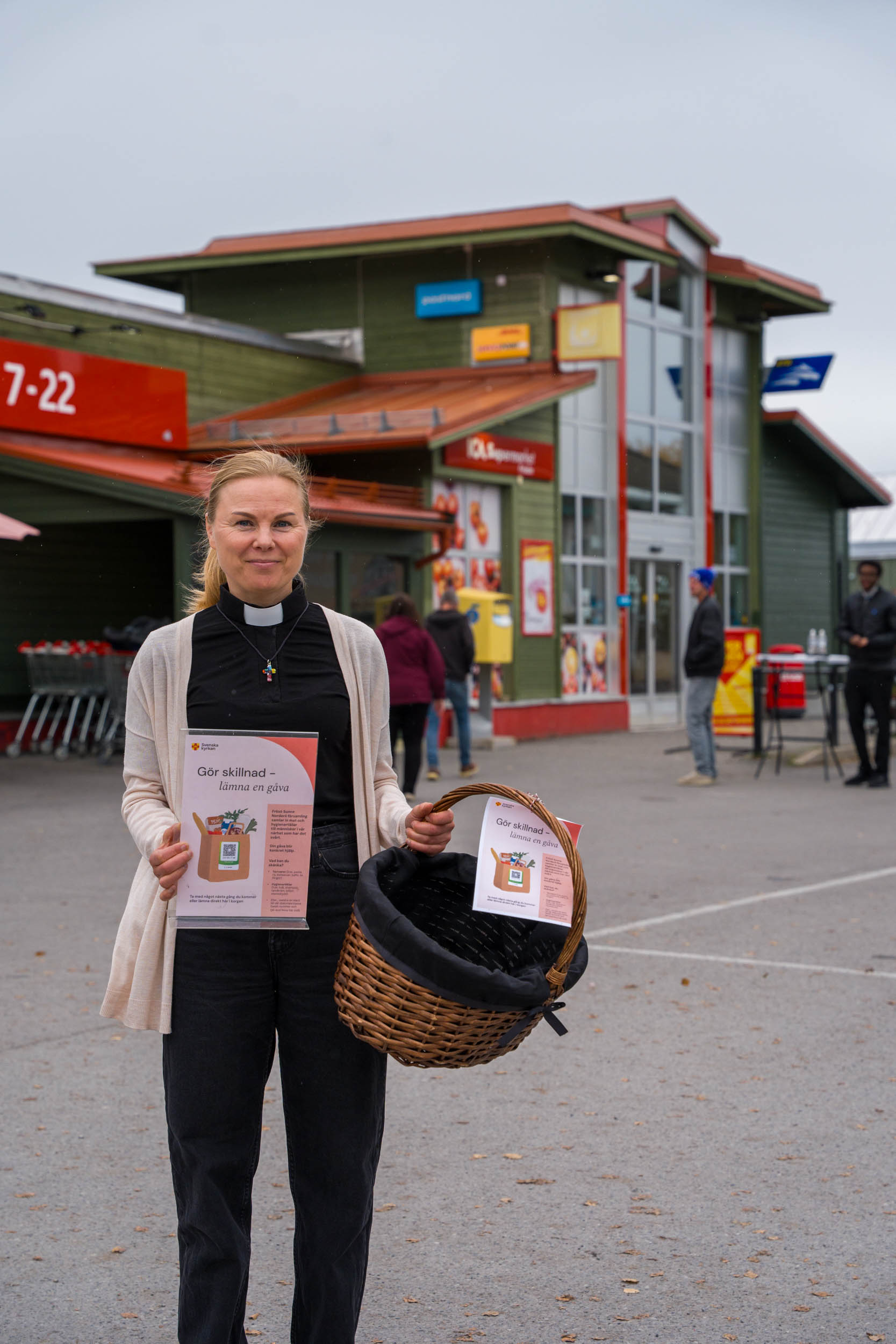 Prästen Johanna står med en matkorg utanför en livsmedelsbutik
