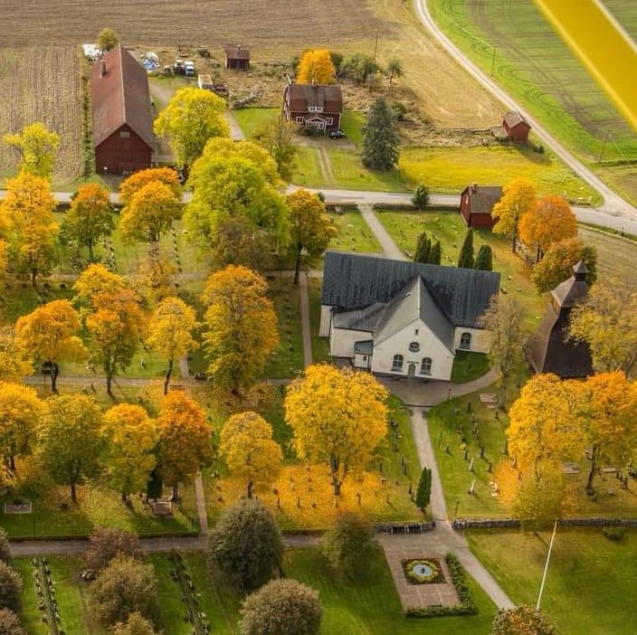 En liten vit kyrka bland fält och höstträd i varma färger och några mindre faluröda byggnader.
