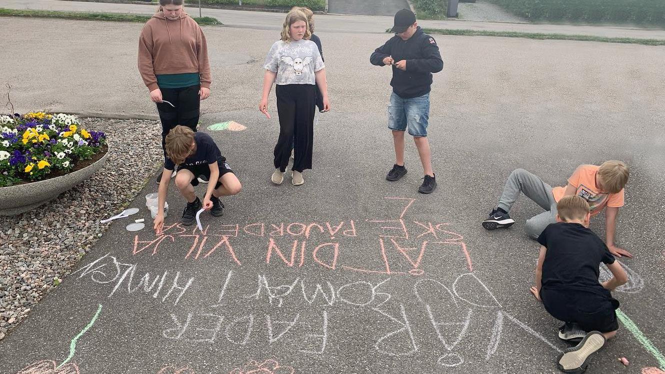 En grupp barn skriver på asfalt: Vår Fader, du som är i himlen.