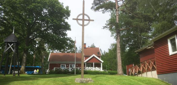 Högt kors på grön gräsmatta med röda byggnader i bakgrunden.