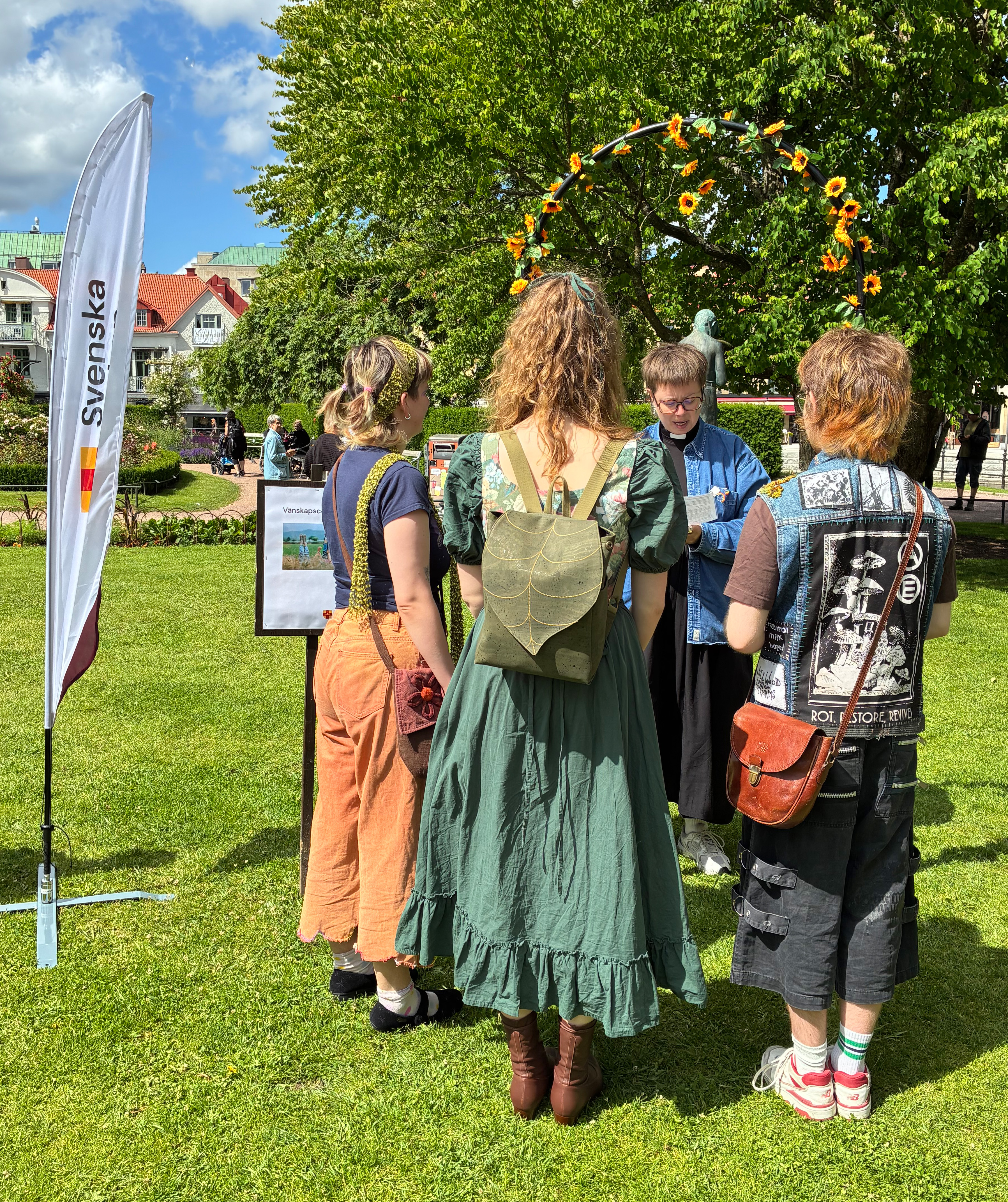 Präst läser för tre unga kvinnor en solig dag i parkmiljö.