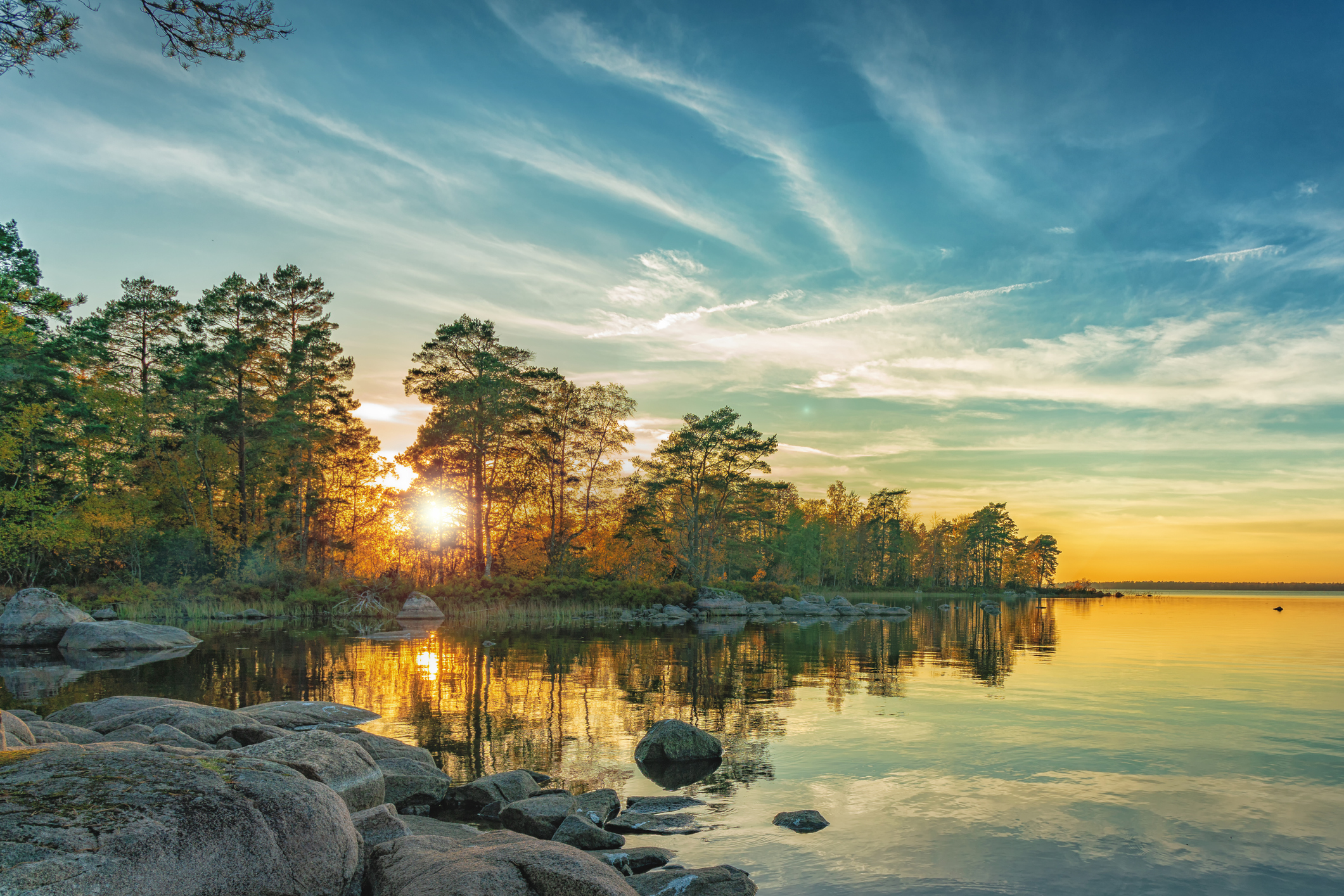 Solnedgång över en stilla sjö med klippig strand i förgrunden. Tallskog kantar vattnet och solen lyser genom träden.