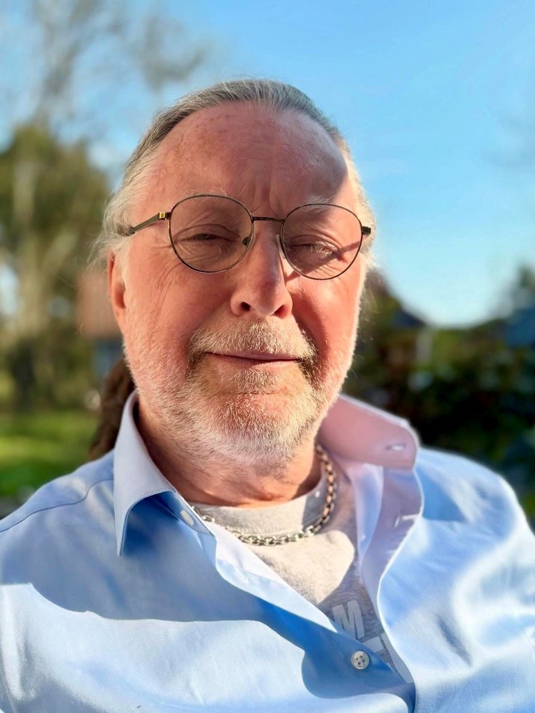 Dan Eiderfors, en 65-årig man kisar lite mot solen i en närbild. Han ler lite. Det ljusa håret är i hästsvans och det vita skägget är kort ansat. Han har ljusblå skjorta, öppen i halsen så en tjock silverkedja syns, runda tunna glasögon. 