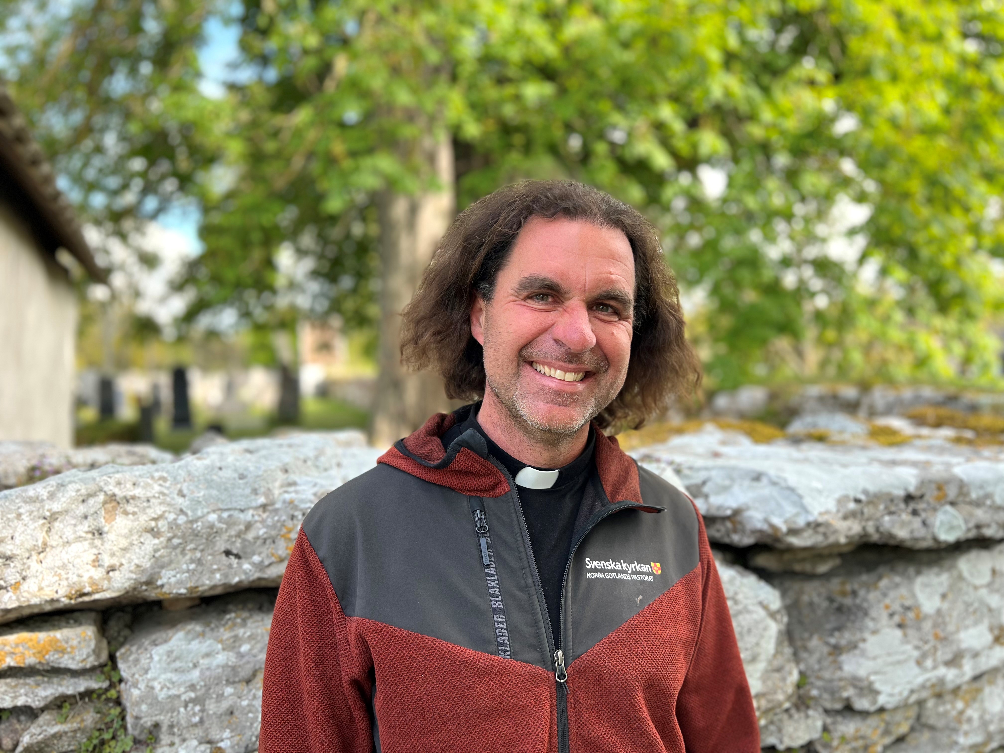 Prästen Johan Sunnerstam står framför en gammal stenmur. Träd grönskar i bakgrunden. Johan ler stort, hans mörka lockiga hår rör sig i vinden. Han har svart prästskjorta och en fleecetröja. 