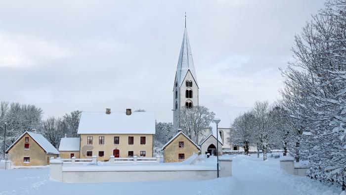 Othem prästgård och kyrka från söder. Prästgården är ljusgul. Kyrkans torn sticker upp högt. Snön ligger mjukt på marken. 