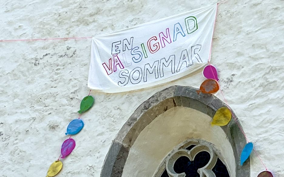 På ett kyrktorn hänger en lakansbanderoll med texten "Välsignade sommar" och ballonger fladdrar i vinden
