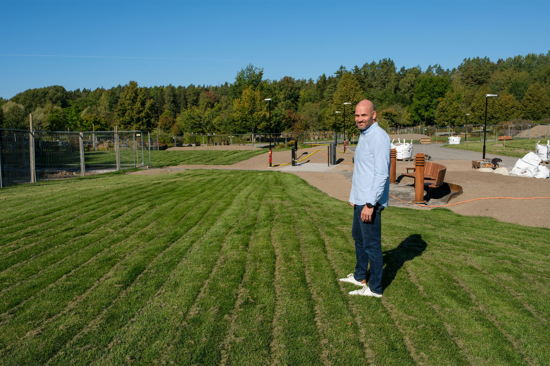 En man i blåa kläder står på en nylagd gräsmatta. Runt gräset syns ett stängsel, vägar och träd