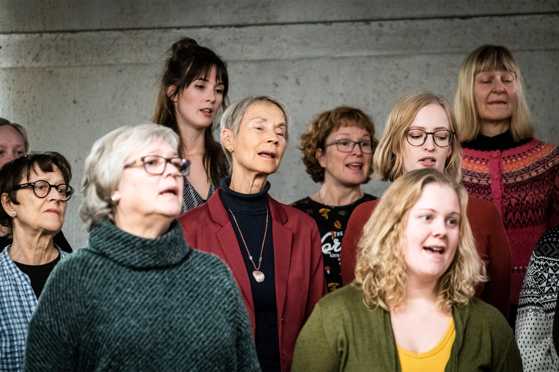 Halvbild på ett tiotal personer som sjunger tillsammans. De står framför en grå betongvägg