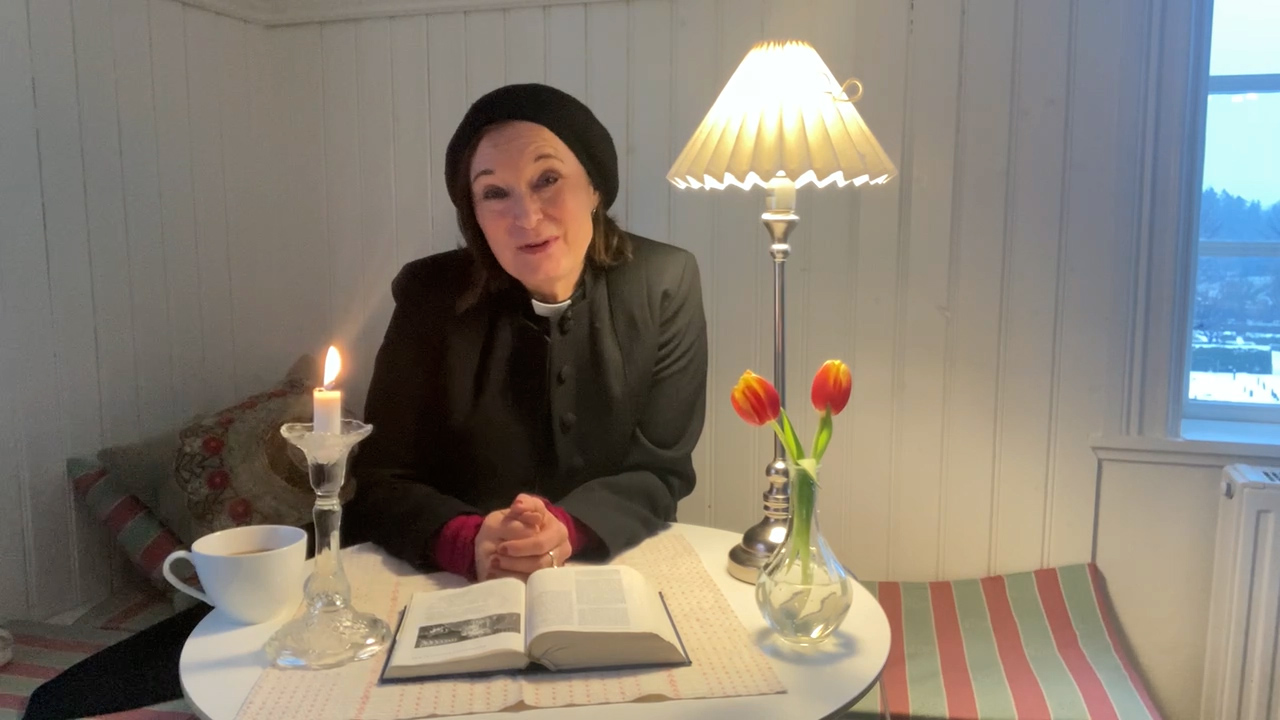 Prästen Anne Sörman sitter vid ett bord med ett tänt ljus, en vas med blommor och en öppen Bibel.